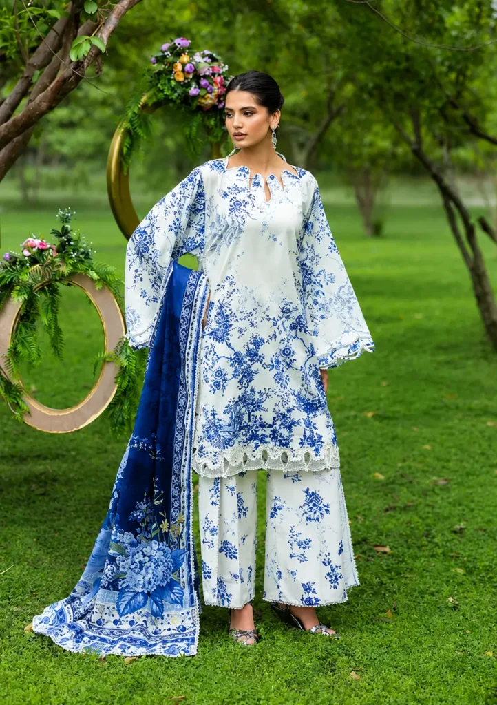 Woman wearing a white and blue printed Pakistani cotton lawn suit with a matching floral dupatta, standing in a lush green garden. The lightweight lawn fabric showcases breathable comfort, elegant prints, and stylish summer fashion.